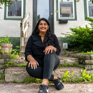 A photo of Aisha Chughtai, a woman with brown skin and long, black hair, wearing a black shirt and slacks, smiling and sitting on the concrete stairs in front of a white house