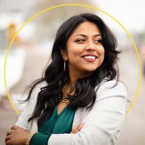 A photo of Aurin Chowdhury, a Bengali American woman with long dark hair, wearing a white blazer and green shirt