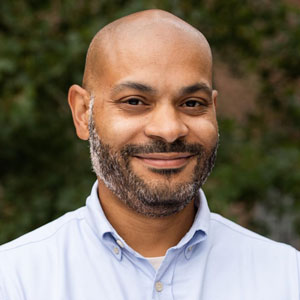 Photo of Elliott Payne, a Black man with a short, dark beard and bald head, wearing a light blue shirt and standing in front of an ivy-covered wall