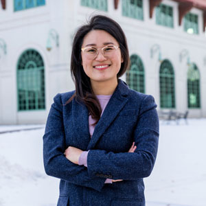 A photo of Hwa Jeong Kim, a Korean-American woman with gold-rimmed glasses and straight, black hair, wearing a blue blazer over a purple shirt