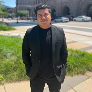 Photo of Jason Chavez, a Mexican American man with short, black hair, wearing a black blazer and tshirt and standing across the street from a stone building in the background