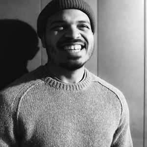 Black and white photo of Jeremiah Ellison, a Black man with a mustache and beard, wearing a knit beanie and sweater, smiling widely at the camera