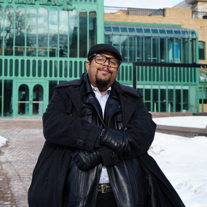 Photo of Marcus Mills, a Black man with a goatee and black glasses, wearing a black hat and coat, sitting in front of a building with a teal metal exterior