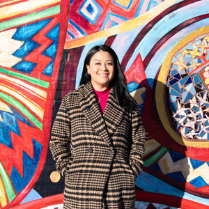Photo of Mitra Jalali, a brown-skinned woman with straight, black hair, wearing a pink shirt under a tweed jacket and standing in front of a multi-colored mural with mosaic glass