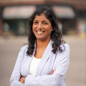 Photo of Saura Jost, a brown-skinned woman with long, curly black hair, wearing a light purple blazer over a white shirt