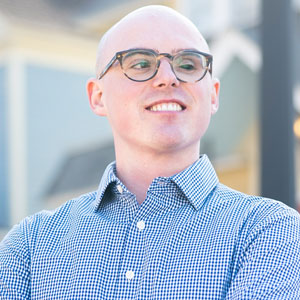 Photo of Soren Stevenson, a white man with a bald head and dark brown glasses, wearing a light blue shirt and standing with his arms folded in front of a yellow and blue house