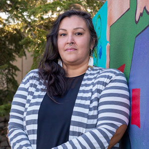 A photo of Miranda Pacheco, an Ojibwe woman with long dark hair standing in front of a painted mural