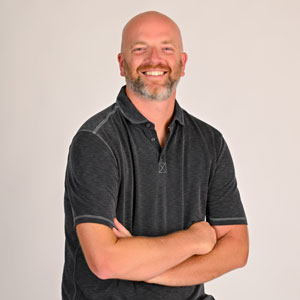 A bald white man with a mustache and short beard wearing a gray polo shirt with his arms crossed