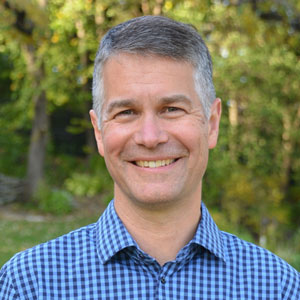 A white man with short gray hair wearing a checked blue collared shirt, smiling in front of a background of trees