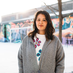 A women with long dark hair, wearing a gray jacket standing in front of a colorful mural in the snow